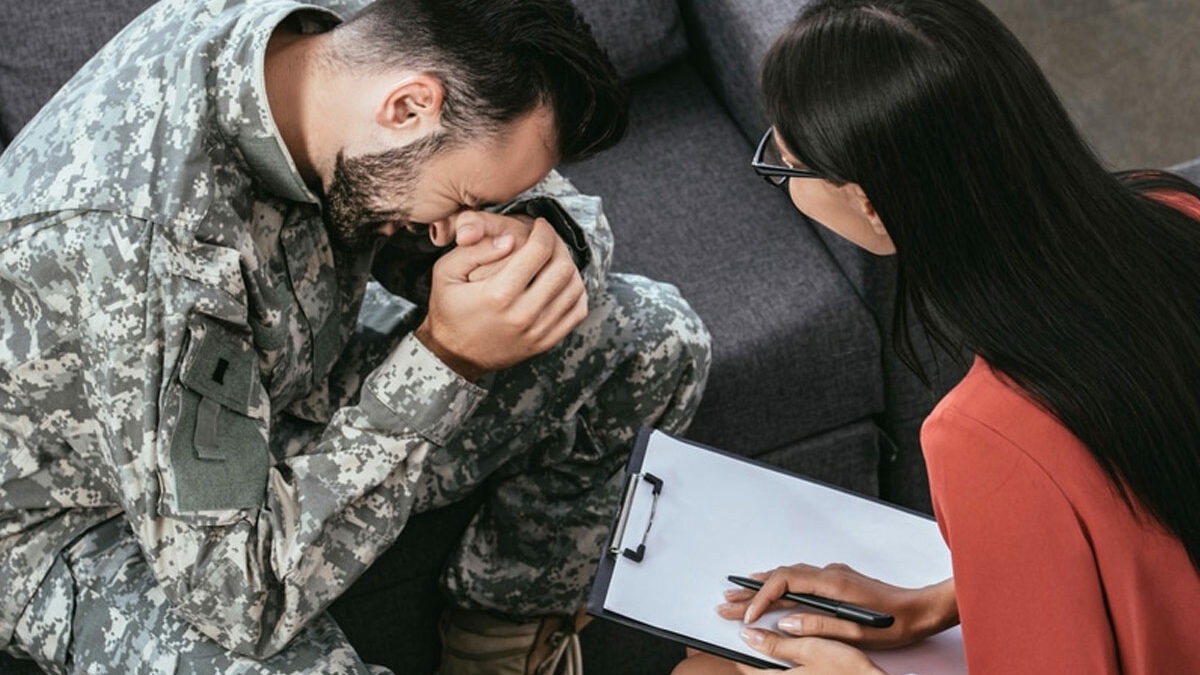 Man in army fatigues, crying with a therapist. PTSD can be triggered by distressing events like accidents, violence, disasters, and loss.